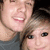 Two people in a close-up selfie with one person's head slightly tilted, wearing a gray shirt, against a dark background.