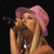A woman with long blonde hair wears a pink bucket hat and black crop top while singing into a microphone against a dark background.