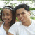 Two people in white shirts stand side-by-side with green foliage background, both smiling at camera.