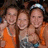 Three people pose together in a static image; two wear orange tops, one wears a light-colored top with dark patterns, and one person wears a silver headband.