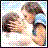 Two people in a close embrace with one person holding the other's face, set against a light blue background with small white dots.