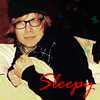 A person with light brown hair and glasses lies on a light-colored surface, wearing a black top with white text and red cursive text reading 'Sleepy' overlaid on the image.