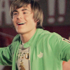 A person in a green zip-up hoodie over a graphic t-shirt with a golden trophy design, smiling with mouth open, against a blurred indoor background.