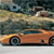 A pixelated orange sports car with dark wheels and blue accents is positioned on a road, with blurred background elements suggesting motion.