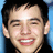 Pixelated close-up of a person with dark hair, light skin, and visible teeth in a wide smile against a blue background.