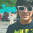 A pixelated image of a person wearing sunglasses and a dark shirt with yellow text, standing outdoors near greenery and a white structure.