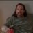 Pixelated static image of a person with long dark hair wearing a dark green top and red lower garment against a muted background.