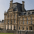 Pixelated image of a historic stone building with multiple chimneys and arched windows under a light blue sky.