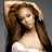 A woman with long brown hair in a white strapless top, hand raised to her head against a gray background.