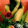 A person in a black top and striped shoes sits with legs drawn up, holding a red rose against a warm-toned background.