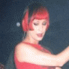 A person with short red hair in a sleeveless red dress appears in multiple poses holding a microphone, wiping face with cloth, and gesturing against dark background.