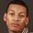 Pixelated close-up of a person's face with short dark hair, light skin, and a checkered shirt collar visible against a dark background.