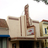 Pixelated static image of a building facade with beige and brown tones, featuring a tall central tower and a small red emblem on a lower section.