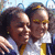 Two people in close proximity, one with dark curly hair and the other with short light hair, both wearing white shirts with yellow accents, against a blurred background.