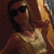 A person in a light-colored tank top and dark sunglasses poses with one hand near their face, captured in a low-resolution, pixelated image with warm brown and orange tones.