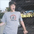 Pixelated image of a person in light gray shirt with circular emblem, dark hair, and blurred background elements.