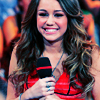 Woman with long wavy brown hair holding a black microphone with red stripe, wearing red sleeveless top and silver necklace, smiling at camera.