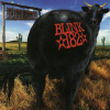 A black cow with red text on its side stands in a desert landscape with cacti and a signpost under a blue sky.