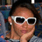 A pixelated close-up of a person with brown hair wearing white-framed sunglasses and a light-colored top, resting their chin on their hand against a blurred background.