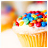 A cupcake with white frosting topped with colorful round candies in red, blue, green, yellow, and orange, set against a blurred background.