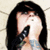 A person with long dark hair and a tattooed neck holds an object near their mouth against a light patterned background.