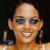 A pixelated close-up of a person with a bald head, dark sunglasses, and a wide smile showing teeth, wearing a light-colored top against a dark background.
