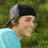 A person in a light blue shirt and black-and-gray cap smiles against a green background, with pink text appearing over the face.