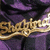 Golden cursive text reading "Shakira" on a purple and black fabric background with visible texture.
