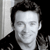 Pixelated grayscale image of a person with short dark hair, wearing a dark jacket over a light shirt, positioned against a light and dark background.
