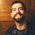 Pixelated portrait of a person with short dark hair and facial hair, wearing a dark shirt against a warm-toned wooden background.