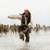 Pixelated figure in dark jacket and light pants extends arm forward against blurred background.