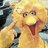 Pixelated yellow bird with orange beak and red tongue sticking out, wearing dark blue clothing against a blurred background.