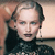 Pixelated close-up of a person with light hair styled back, wearing a dark high-necked top against a blurred background with warm tones.