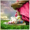 A person in a pink garment bends over a colorful sneaker on green grass under a cloudy sky.