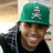 A person wearing a green cap with a white skull and crossbones symbol, dark hoodie, and black shirt; the right side shows green text overlay.