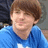 Pixelated image of a person with brown hair wearing a blue shirt, cropped to show upper body against a blurred background.