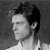 Black-and-white pixelated image of a person's head and shoulders in profile view, facing left, with short dark hair and a light-colored shirt against a dark background.