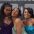 Three people in a group pose wearing sleeveless dresses, one purple, one light green, and one blue, against a dark background with vertical lines.