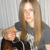 A pixelated image of a person with long light brown hair wearing a dark gray shirt with a white pattern, holding a brown object against a light gray striped background.