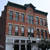 Pixelated static image of a multi-story brick building with dark roof, gray stone base, and multiple windows under a light sky.