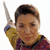 A person with short brown hair wearing a yellow and red plaid shirt holds a silver object above their head against a white background.