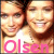 Two people with light brown hair and fair skin appear in a pixelated close-up, wearing white tops against a warm-toned background with pink text below.
