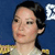 Pixelated static image of a person with dark hair tied back, wearing a light-colored top with gold shoulder accents against a blue background.