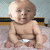 A baby in a diaper sits on a patterned surface with a light blue wall and dark trim in the background, showing multiple facial expressions and holding an orange object.