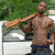 A shirtless man with dark skin stands outdoors holding a long wooden object over his shoulder, positioned in front of a white vehicle and green foliage.