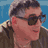 Pixelated static image of a person with short dark hair wearing sunglasses and a brown jacket against a blue background.