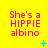 Yellow background with pink text reading "She's a HIPPIE albino" repeated in a , each containing a small blue plus or star symbol