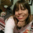 A person with shoulder-length brown hair and bangs wears a white long-sleeve shirt under a red-and-white striped sweater, resting their chin on their hand with a smile.