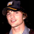 Pixelated static image of a person wearing a dark cap with yellow trim and light-colored shirt against black background