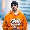 A person wearing an orange hoodie with a white and black graphic logo, a dark beanie, and a white undershirt, standing against a blurred background.